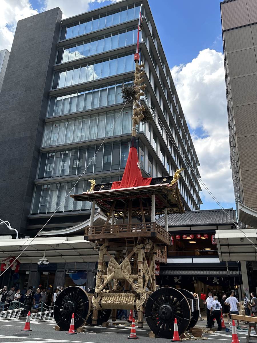 祇園祭的鉾車停在京都市街,車體下半部完全以粗繩綑紮成交叉花紋,兩側是黑色大車輪,頂端真木高過旁邊的辦公大樓