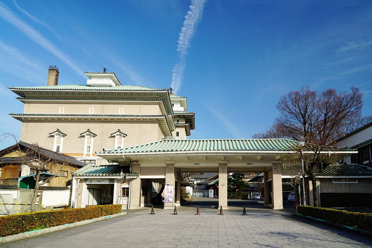 晴天下的祇園甲部歌舞練場:後方是綠色琉璃瓦屋頂、米色外牆的多層樓大屋頂建築,頂上有一座望樓;前方是同樣綠瓦的車寄門廊,柱間拉了三角錐,廣場上沒有人。