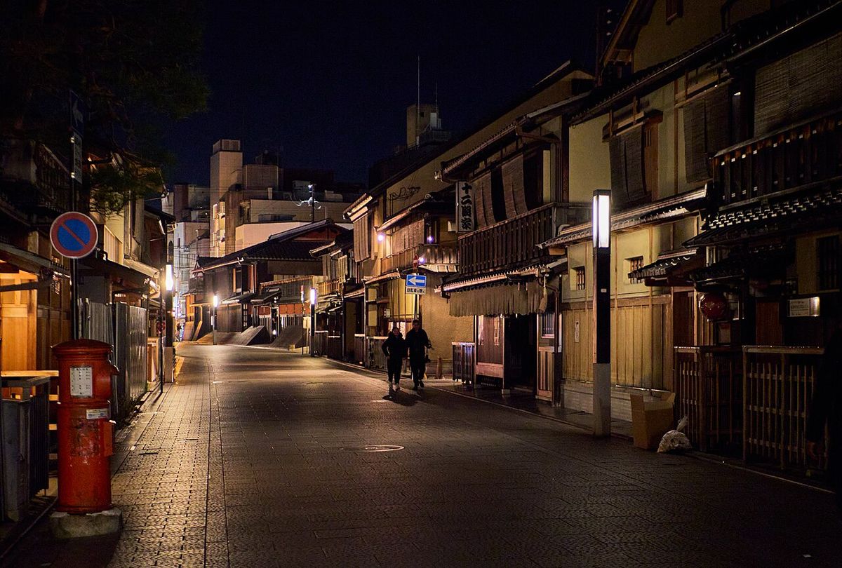 京都祇園夜間的石板路街道,兩側是二層樓木造町家,右側屋簷下垂著長暖簾、旁邊立著白色柱燈,左前方是紅色圓筒郵筒與禁止停車標誌,街道中段兩名行人背對鏡頭走遠