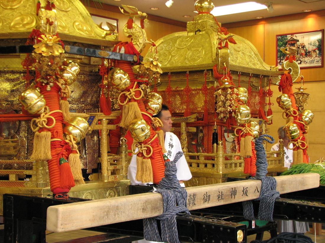 三基金色神輿並排停放在室內,紅繩纏繞的柱上掛滿金鈴,前方橫木刻著「八坂神社御神輿」