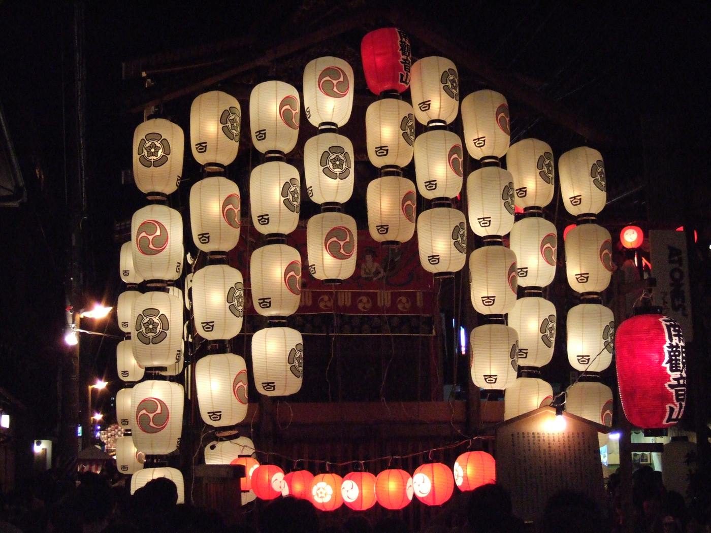祇園祭宵山夜晚:山鉾上一整面點亮的白色駒形提灯,下方串著一排紅色小燈籠,畫面最下緣是人群的頭部剪影
