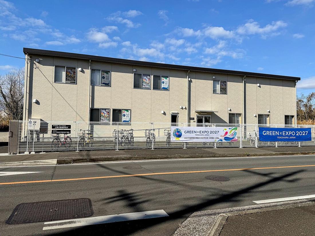 橫濱市脫碳・GREEN×EXPO 推進局上瀬谷整備事務所的兩層米色組合屋,圍籬上掛著白色與藍色兩幅「GREEN×EXPO 2027 YOKOHAMA JAPAN」布條,窗內貼著博覽會海報