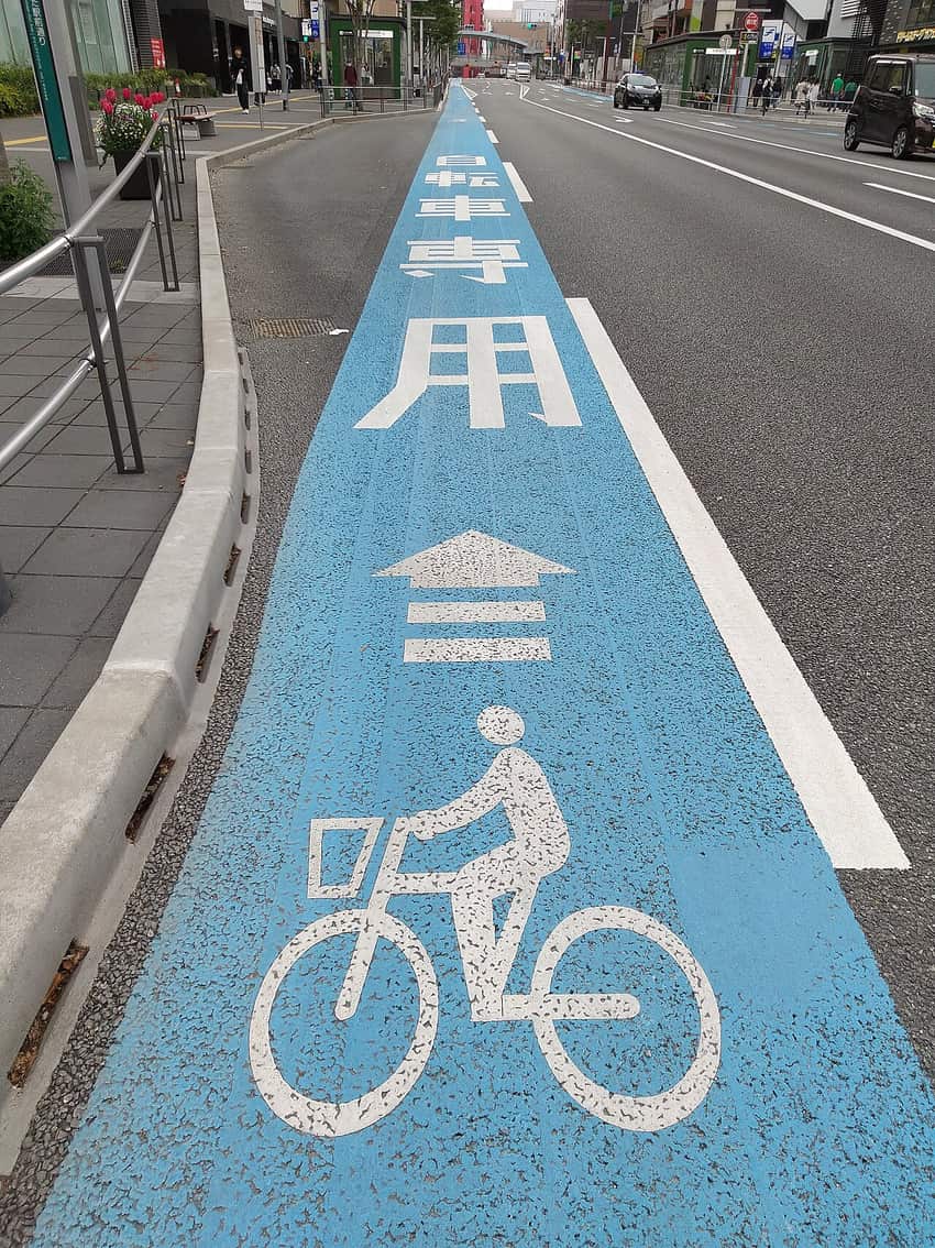 福岡博多站前大道:車道最左側一整條漆成水藍色,路面白字寫著「自転車専用」,下方接一支三道人字紋的前進箭頭與一個大型單車圖示,右邊以白色實線與汽車車道分開,左邊是水泥分隔塊與人行道,遠處對向車道有車輛車頭朝鏡頭