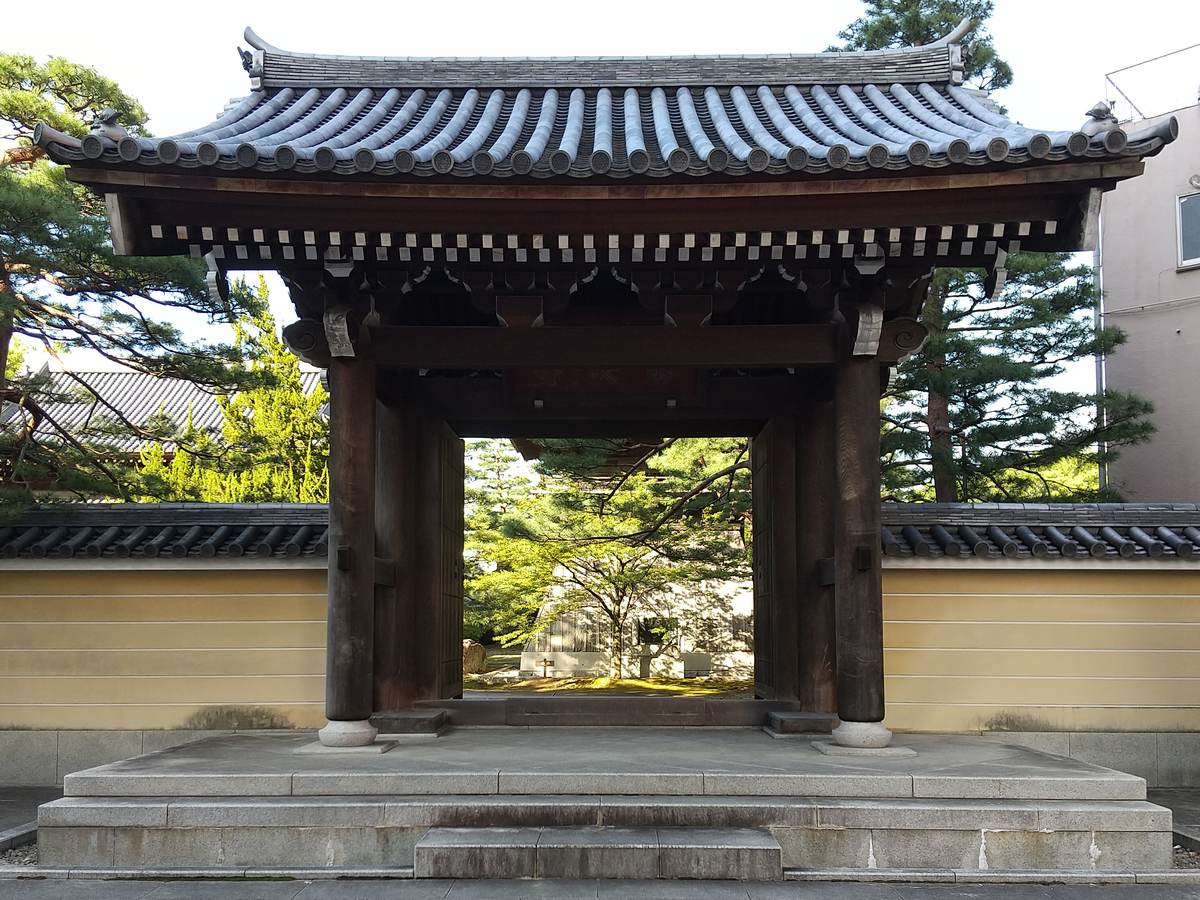 承天寺的木造山門,深色梁柱與黑瓦屋頂立在花崗岩台座上,兩側是米黃色土牆與松樹,門扉敞開可以看到院內的樹
