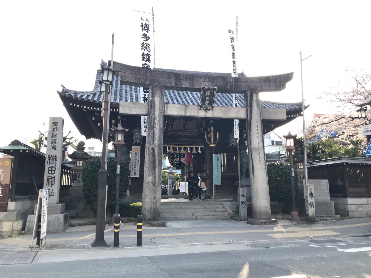 櫛田神社的石造鳥居與後方木造樓門,鳥居中央掛著寫有「櫛田宮」的匾額,兩側白色幟旗寫著「博多総鎮守」,右側一株櫻花正在開