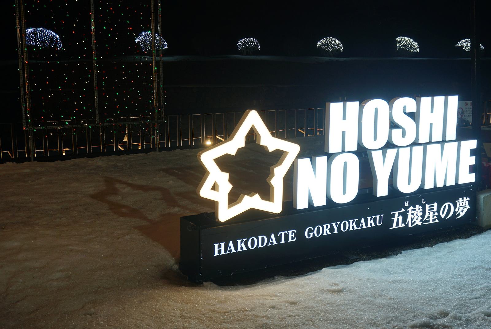 雪地上一座發白光的立體字看板,左邊是星形框,右邊是大字「HOSHI NO YUME」,下方黑底寫著「HAKODATE GORYOKAKU 五稜星の夢」;看板後方是柵欄與護城河對岸的土堤,堤上一排灌木被網狀白燈包成圓球,左側有一座掛滿紅綠小燈的鐵架