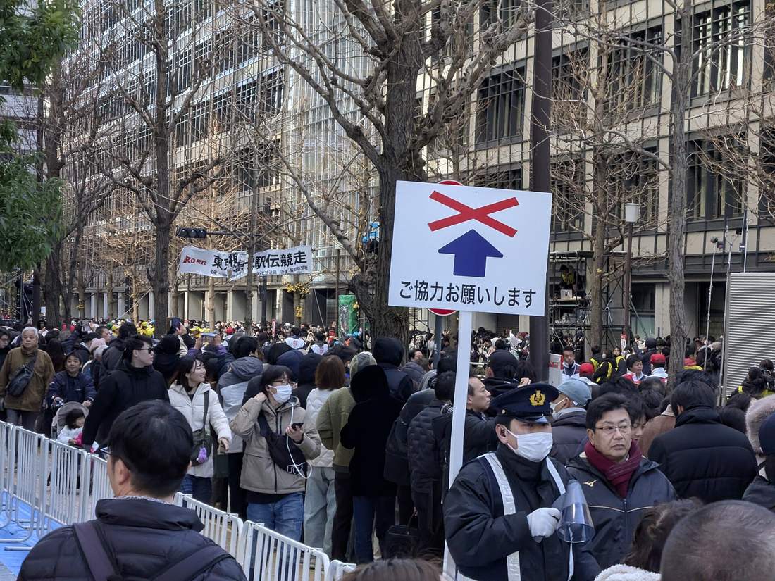辦公大樓夾道的街上擠滿觀眾,人群被白色鐵欄杆隔在人行道上,畫面中央立著一塊白色告示牌,上面是一個被紅色叉叉打掉的藍色箭頭與「ご協力お願いします」字樣,牌子下方一名戴警帽與口罩的警察拿著擴音器,左後方拉著一條寫有「東京箱根間往復大学駅伝競走」的橫幅,行道樹是落葉後的光禿枝條