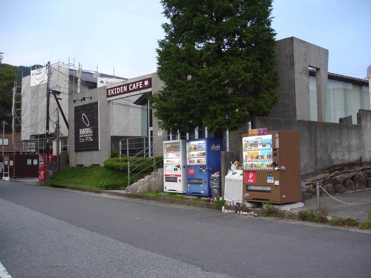 路旁的清水混凝土建築,牆面上掛著深色看板,上面是一圈由多色線條組成的環形標誌與「箱根駅伝ミュージアム」字樣,入口上方另有一塊寫著 EKIDEN CAFE 的招牌,建築左半邊架著鷹架並罩著白色帆布,路邊並排著四台飲料自動販賣機,前方是空無一人的柏油路