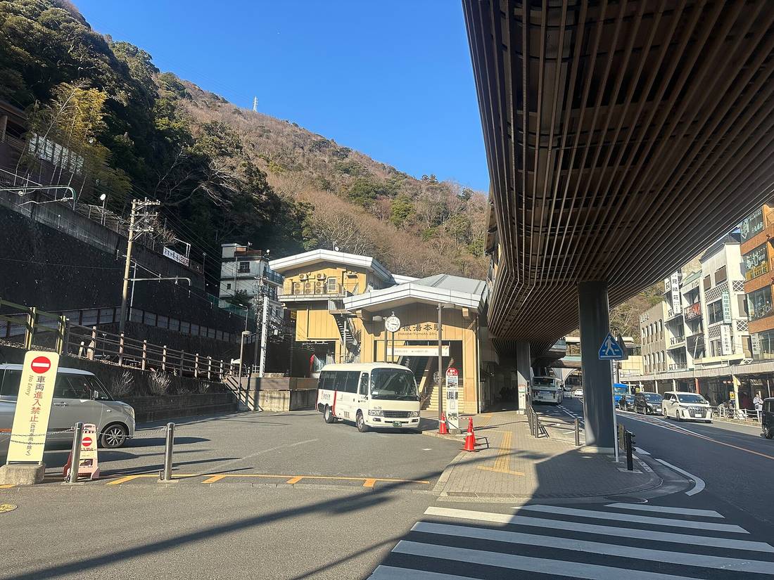冬日晴天的箱根湯本站站前,米黃色的站房正面掛著圓形時鐘與「箱根湯本駅 Hakone-Yumoto Sta.」招牌,站前迴車道上停著一輛白色小型接駁巴士與幾支橘色三角錐,左前方立著黃色的「車両進入禁止 No Entry」立牌,右上方是木格柵構成的人行天橋雨庇,背景是落葉後的褐色山坡與商店街建築