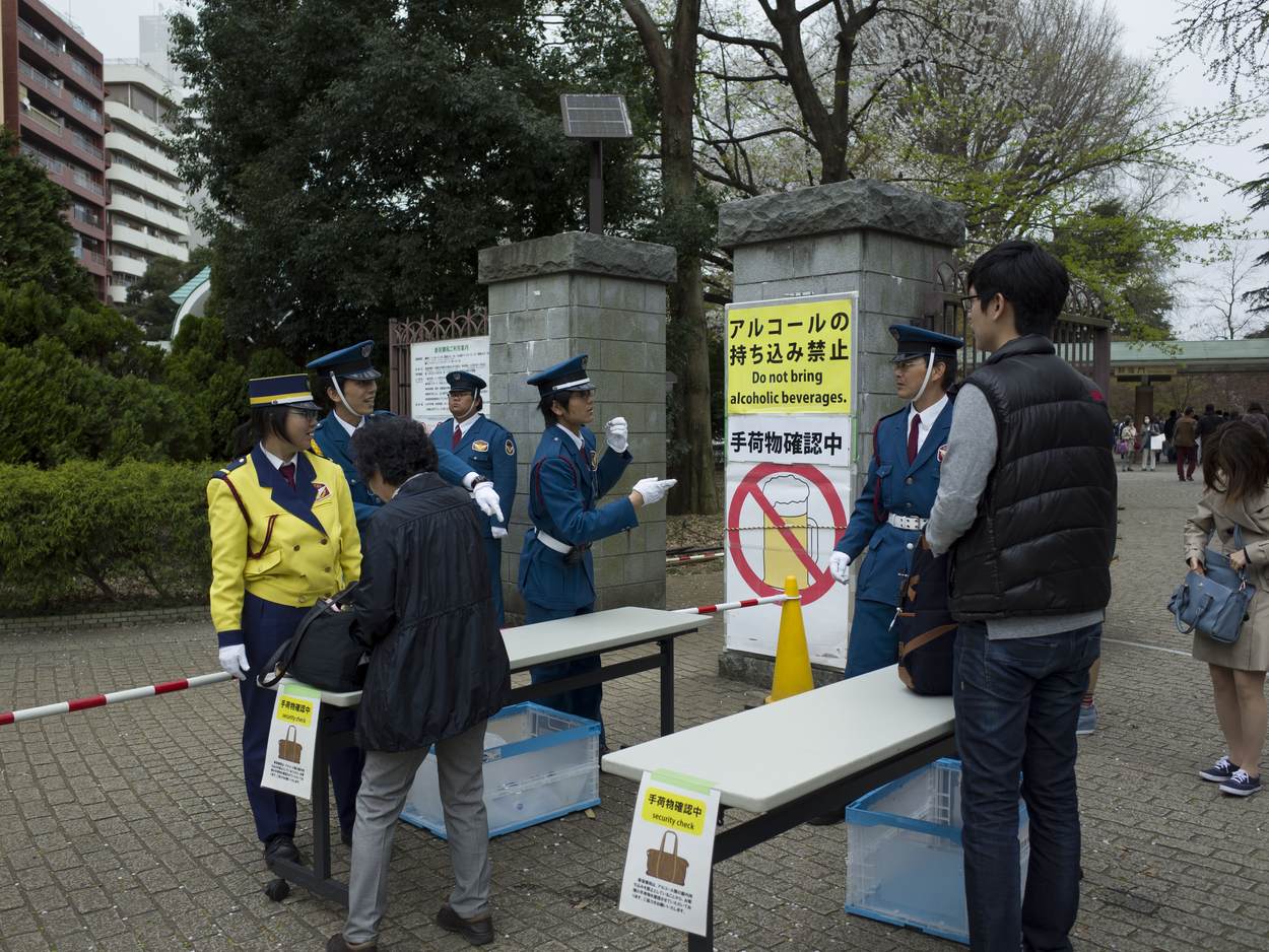 新宿御苑入口設著長桌與塑膠籃檢查手提行李，旁邊立著「禁止攜入酒精 / Do not bring alcoholic beverages」的告示牌與畫叉的啤酒杯圖示，六名警衛在旁引導