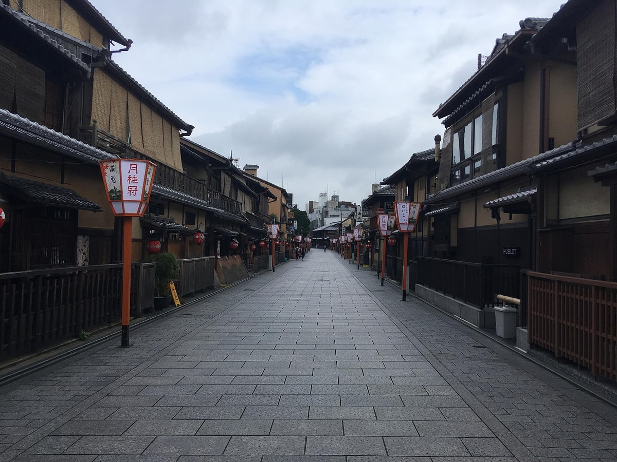 清晨無人的花見小路:整條石板路一路延伸到遠處,兩側是二層木造町家與紅色圓燈籠,路旁一排橘色柱燈上寫著「温習会」與商號名,遠處只有兩個看不清面孔的行人。