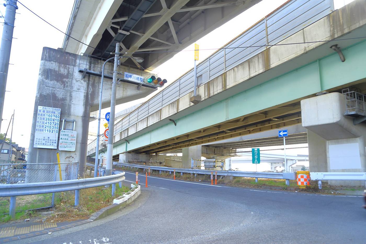 西湘 Bypass 高架橋下的平面道路,頭頂是兩層綠色鋼樑高架橋,左側橋墩上掛著白底料金表看板,列著普通車、大型車、特大車、中型車、軽自動車五段金額,中央號誌燈旁掛著綠色路牌「旧早川橋際」,右側立著一面綠底白字的直式看板寫著「早川インター入口」