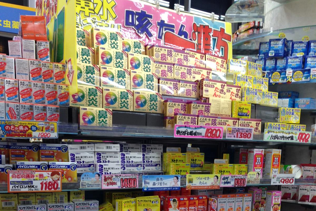 日本藥妝店的成藥貨架,上層整排是印著大字「鼻炎」的藥盒,旁邊接著「咳」「痰」與感冒藥、兒童用感冒藥,每一格都插著粉紅與黃色的日圓價格牌。