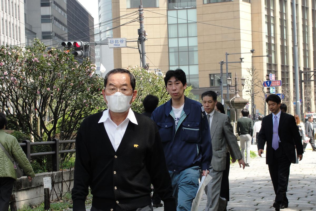 東京日比谷街頭,一名戴眼鏡與白色摺疊口罩的男性走在人行道上,身後幾位沒戴口罩的上班族與工作服男子並行,左側花壇的矮樹開著粉色花,路口掛著「日比谷」標示。
