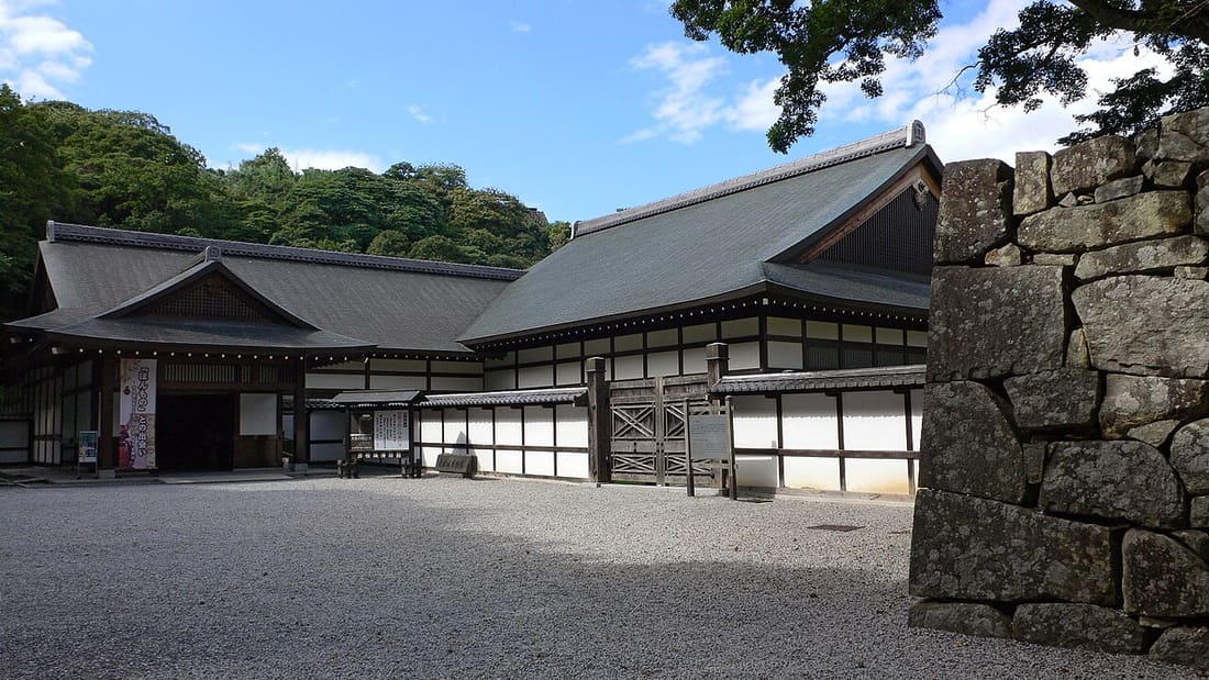 彥根城博物館外觀:兩棟入母屋造的木造建築沿著碎石廣場排開,左側是玄關與掛著直式布幔的入口,中央有白灰泥牆與木格柵的長屋、一道交叉木條的板門,右前方是一段粗石堆砌的石垣