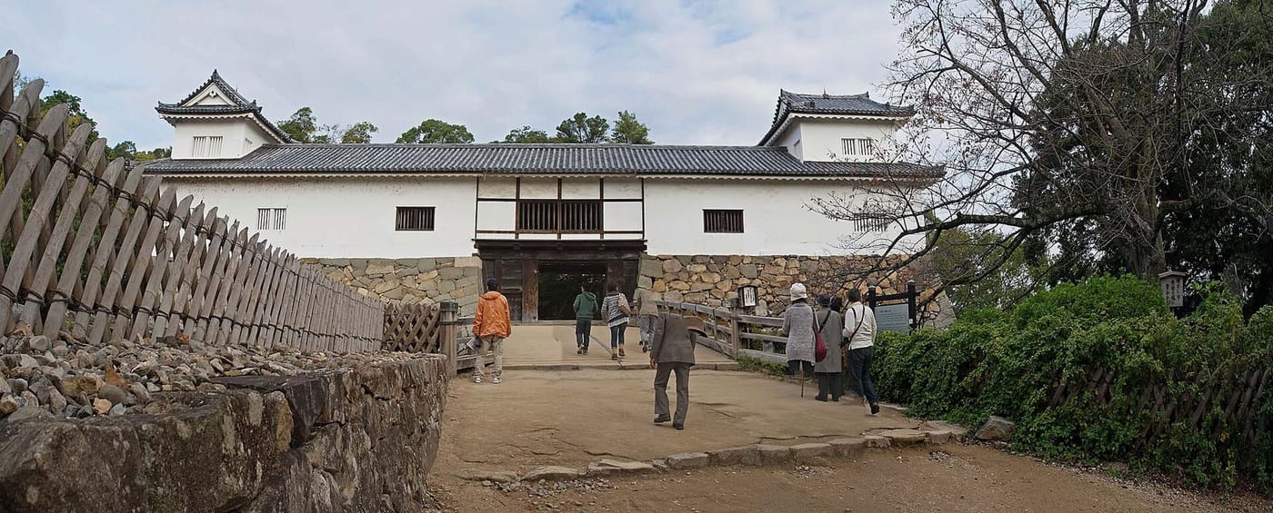 彥根城天秤櫓正面:一棟長條形的白牆建物架在石垣上,兩端各有一座小型的黑瓦屋頂角櫓,正中央下方是一個開著的木造門洞,一座木橋跨過門洞前的空隙,十來名遊客沿著土坡走上橋;左側是斜插的木柵,右側是灌木叢與立牌
