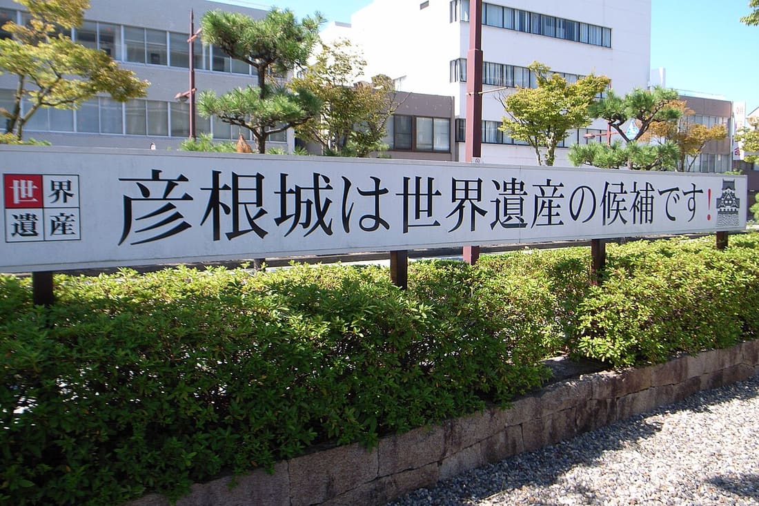 彥根市街道旁一面長條白色看板,黑色大字寫著「彥根城は世界遺産の候補です!」,左端有一個寫著「世界遺產」的方框、右端是天守的小圖示,看板立在修剪過的矮樹籬後方