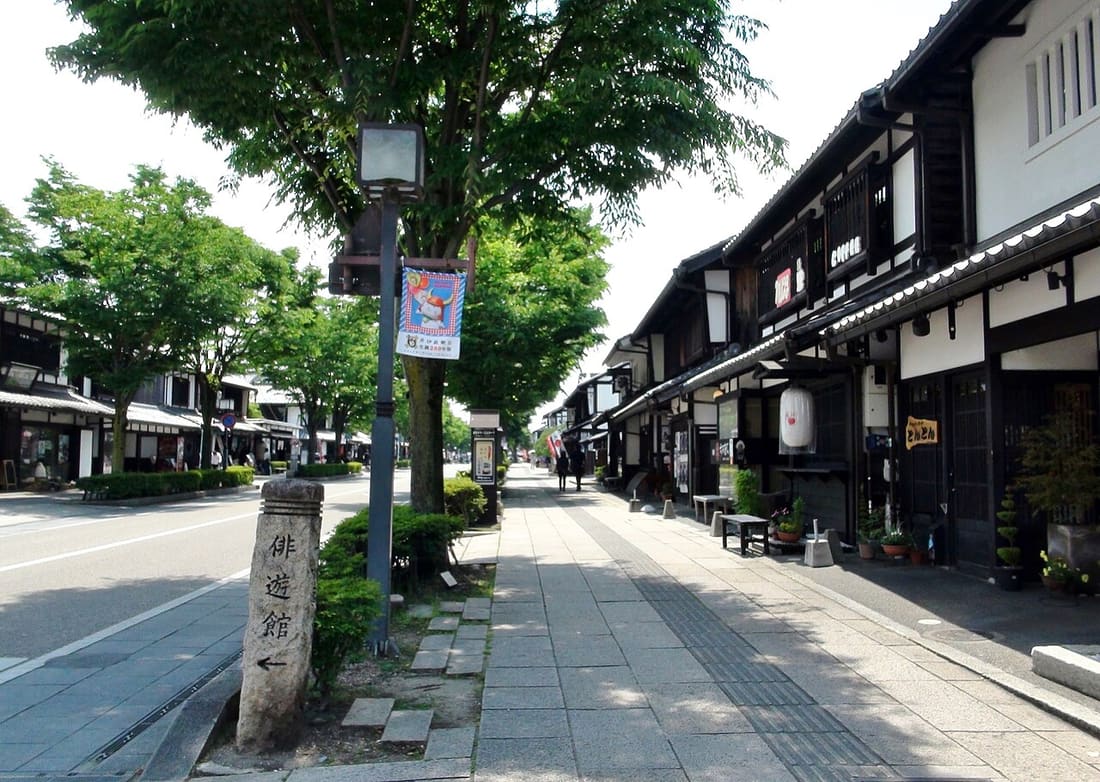 彥根夢京橋城堡大道:白牆黑木格的町家沿街並排、屋簷下掛著燈籠與布招牌,人行道以石板鋪成,左側是行道樹與雙向柏油車道,路燈上掛著一面直式旗幟