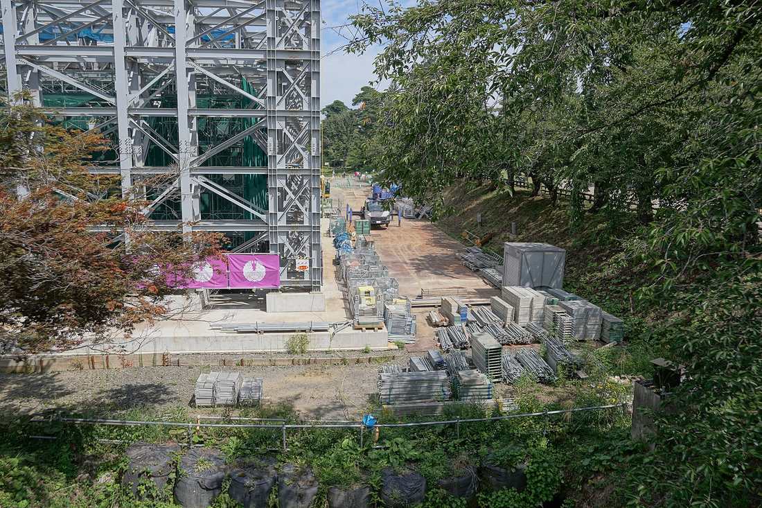 從高處俯瞰弘前城內濠工事現場,灰色鋼構作業構台從乾涸的濠底架起、內側掛滿綠色防護網,構台底下是一片混凝土基座,右側堆著大量鷹架板與鋼管