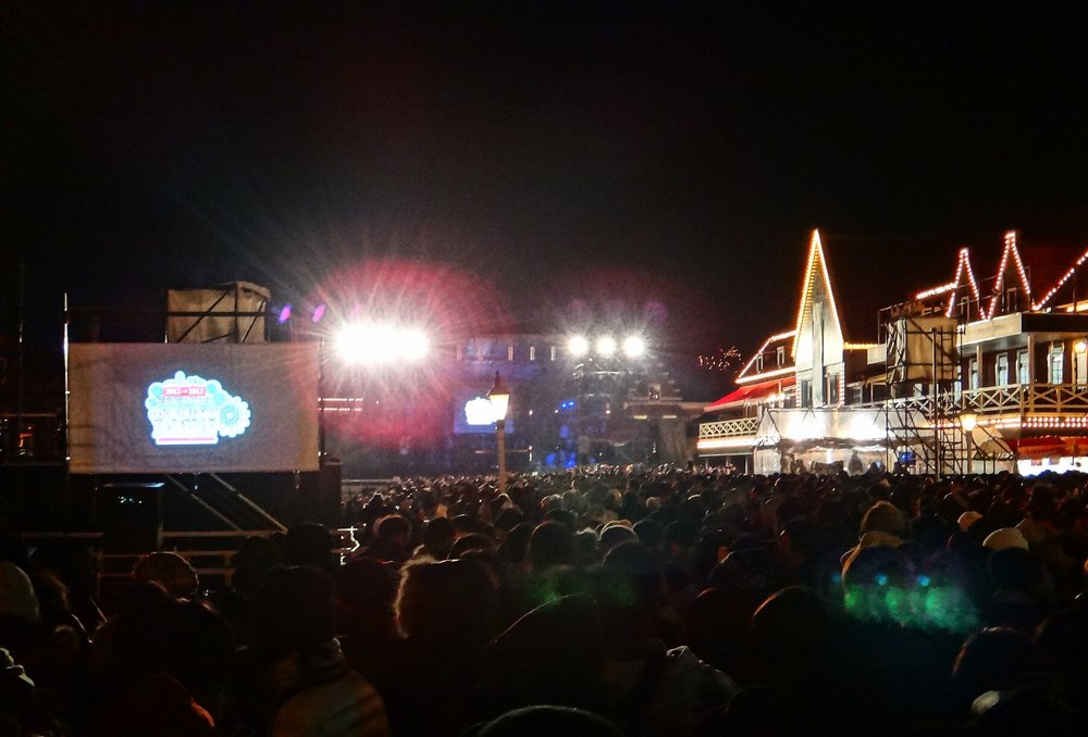 豪斯登堡港區跨年夜的倒數會場:左邊大螢幕顯示 2012→2013 倒數標誌,中間舞台打燈,右邊是掛滿燈串的荷蘭式建築,前方擠滿站著等倒數的人群背影