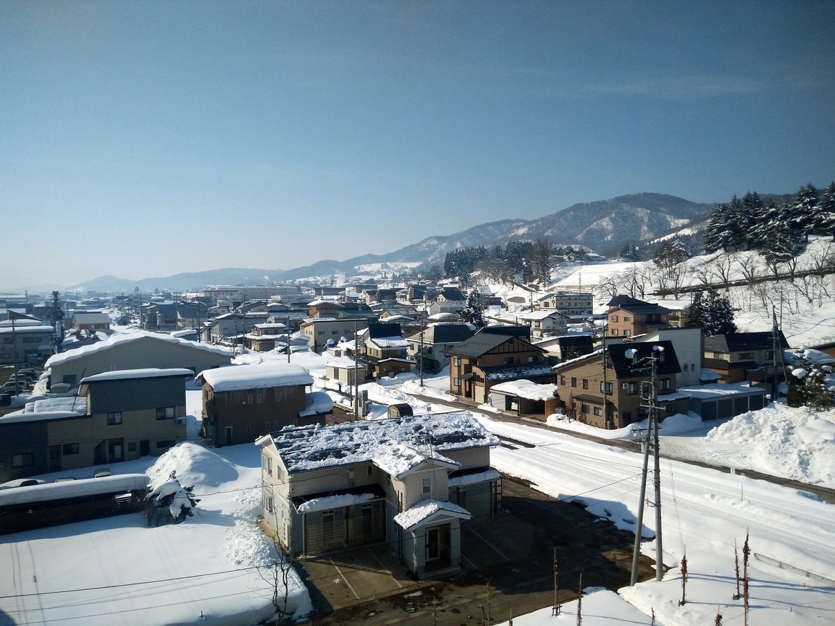 二月晴天從飯山站往外看的飯山市區：住宅屋頂積著厚雪，房子之間堆著剷出來的雪堆，右側那條路的路面整條是壓實的白雪與車轍，遠方是積雪的山稜