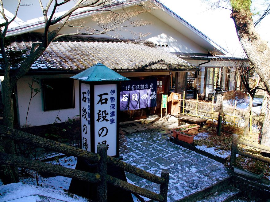 冬季的石段之湯外觀,瓦頂建築前立著印有「伊香保溫泉 石段之湯」的燈箱招牌與同名暖簾,石板地上有殘雪
