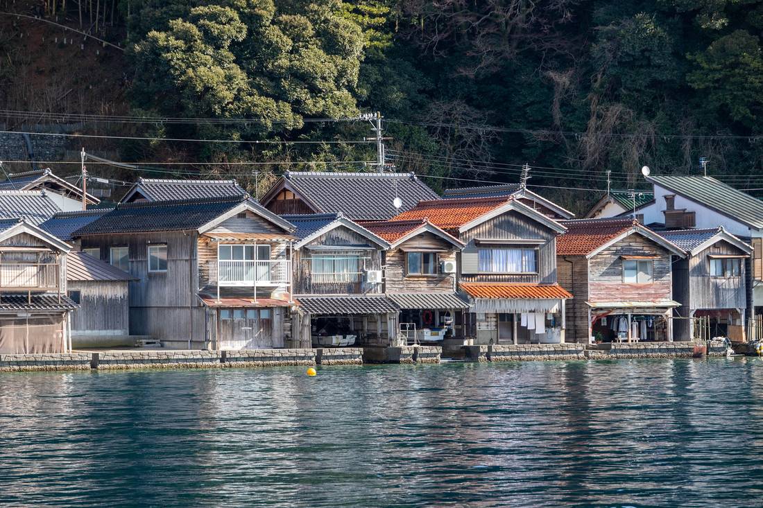 從海面看伊根町的舟屋群:一整排木造房屋沿著岸線並排,一樓的船庫直接開向水面、石砌基座泡在海裡,二樓是有窗有陽台的住居層,屋後是常綠樹覆蓋的陡坡