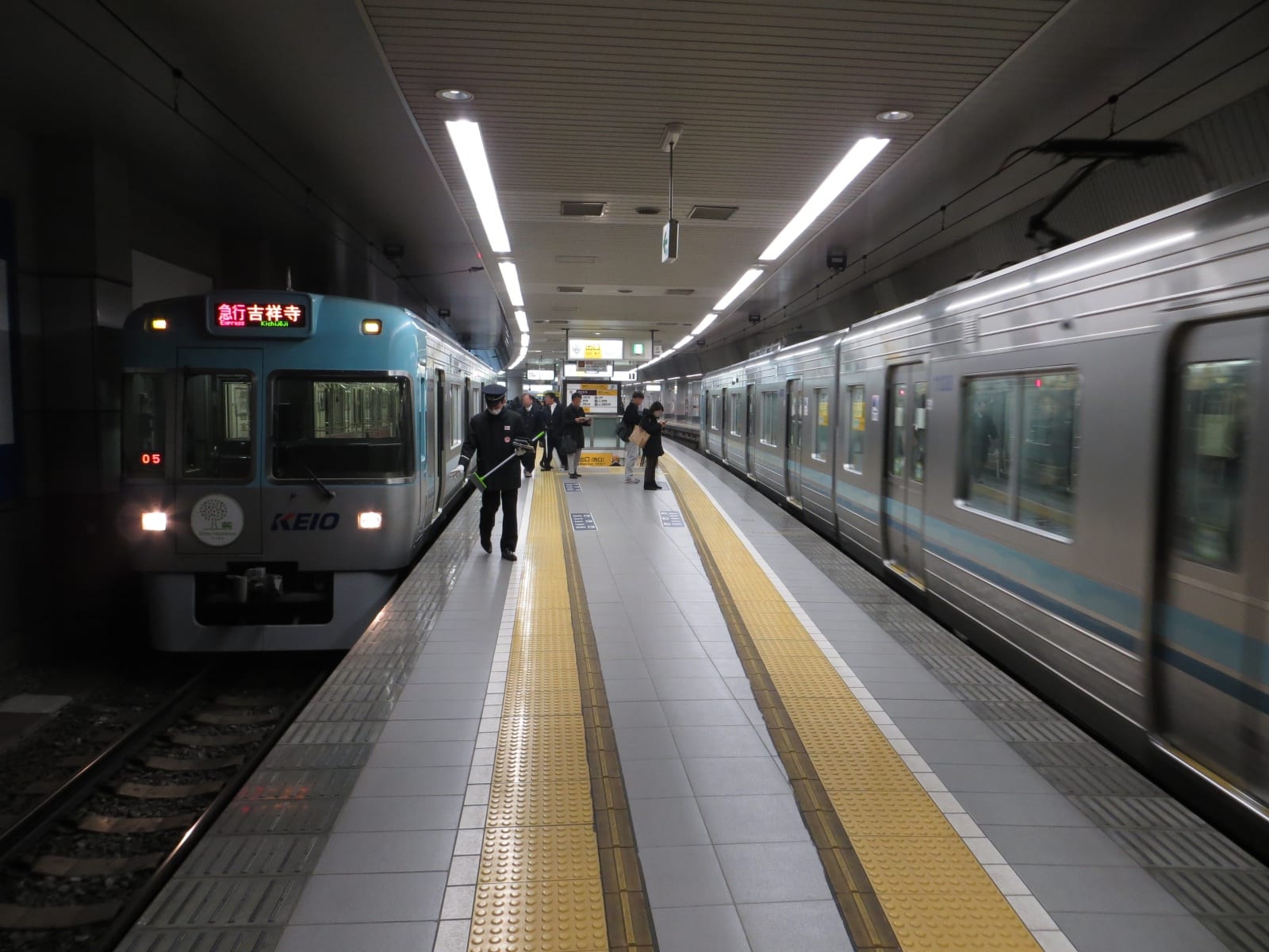 地下月台上停著一列京王電鐵的電車,車頭下方印著 KEIO,上方 LED 以紅字顯示「急行 吉祥寺 Express Kichijōji」,穿制服的站務員拿著長柄工具走在月台中央,右側另一列電車正貼著月台通過