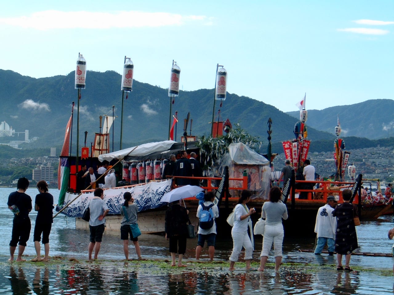 管絃祭御座船停在宮島淺灘上,船上掛滿提燈與幟旗,觀眾赤腳站在水中圍觀