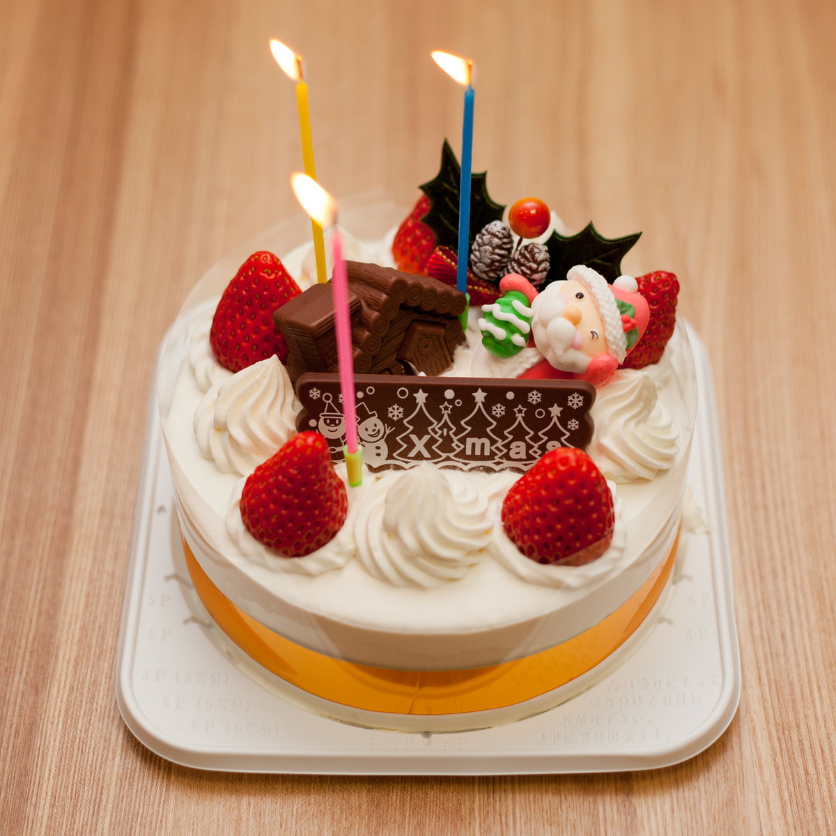 日本聖誕蛋糕：白色鮮奶油圓形海綿蛋糕鋪上整顆草莓，插著巧克力 X'mas 牌、聖誕老人糖偶與三支點燃的蠟燭