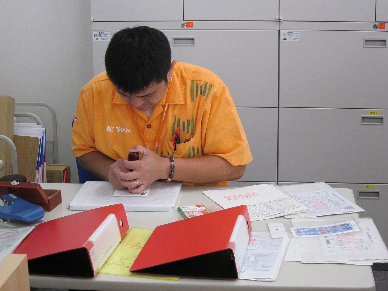 日本郵局櫃檯後方,一名穿橘色制服的男性職員低頭在一份文件上蓋章,制服左胸繡著 JP 標誌與「郵便局」三個字,桌面攤著兩本紅色檔案夾、數份印著紅色格線的表單與一枚印台,身後是整面灰色的文件櫃