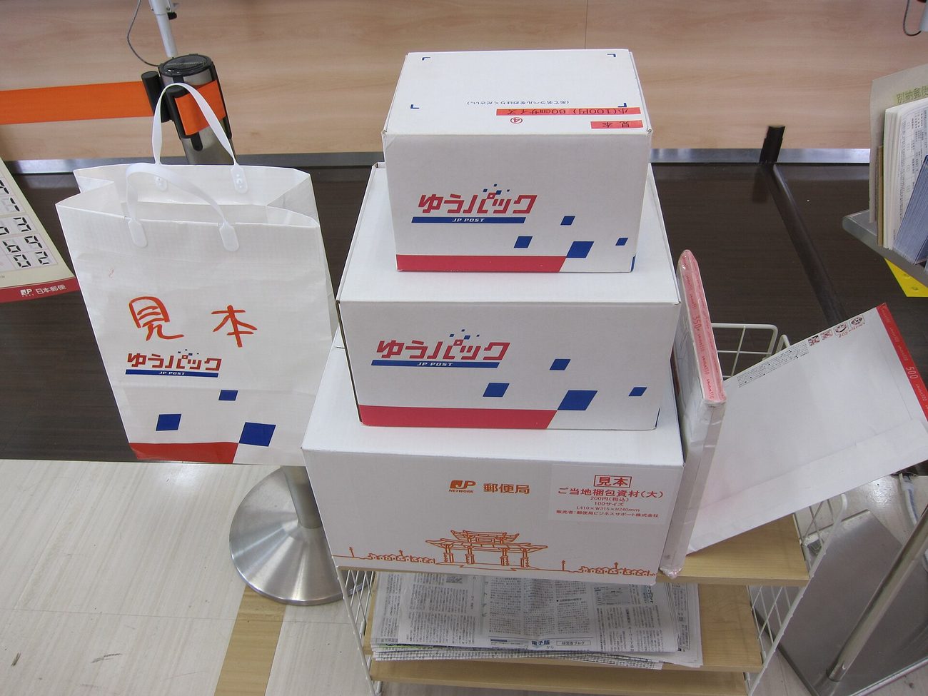 郵局櫃檯旁的樣品架上擺著三件日本郵便包裝材:一個印著紅字「見本」與 ゆうパック JP POST 標誌的白色手提紙袋,兩個白底配紅藍幾何色塊的 ゆうパック 紙箱,最下方的大紙箱印著 郵便局 標誌、橘色線條畫的城門圖案與「ご当地梱包資材(大)200円(税込)」及尺寸 L410×W315×H280mm