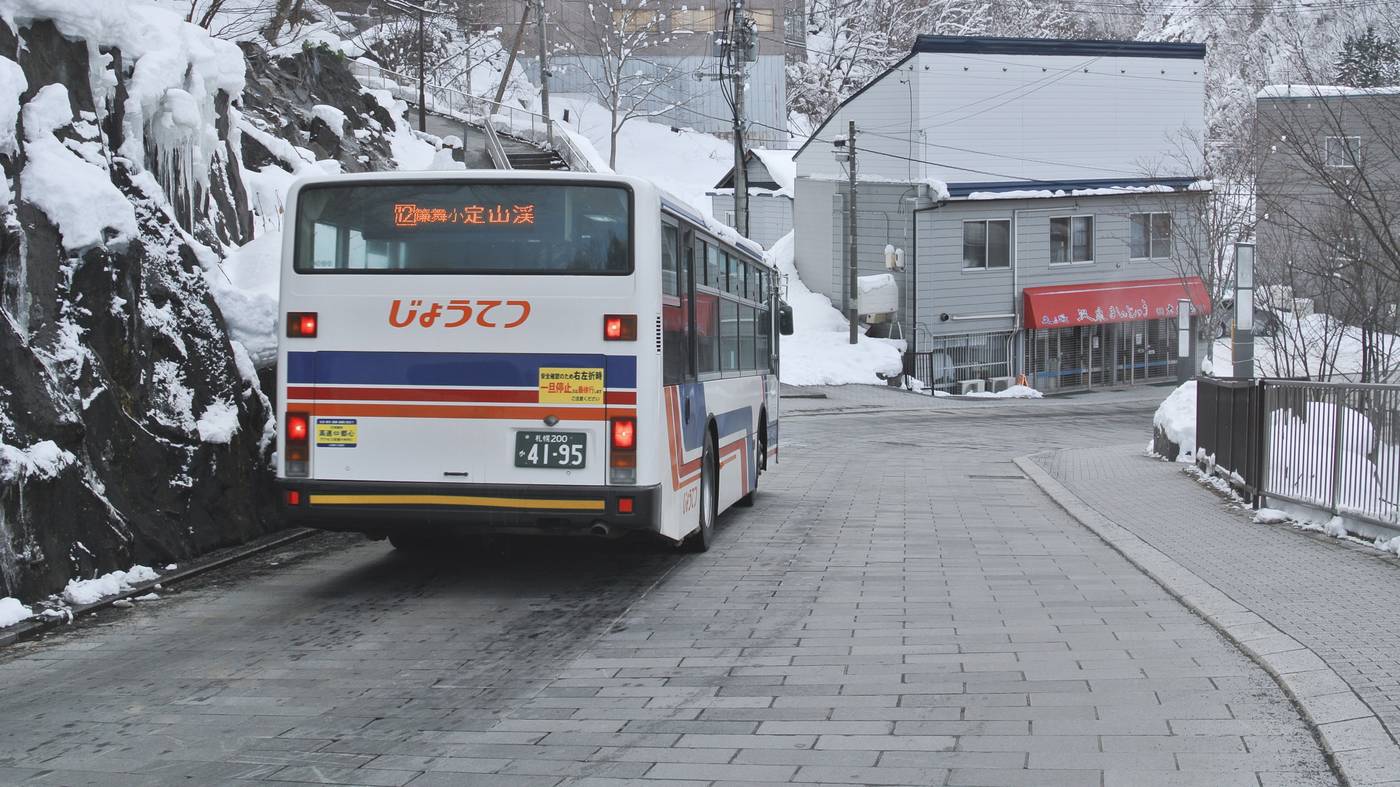 從後方看じょうてつ的路線巴士,車尾看板顯示 12 號系統往定山渓,開在兩旁積雪的石板坡道上