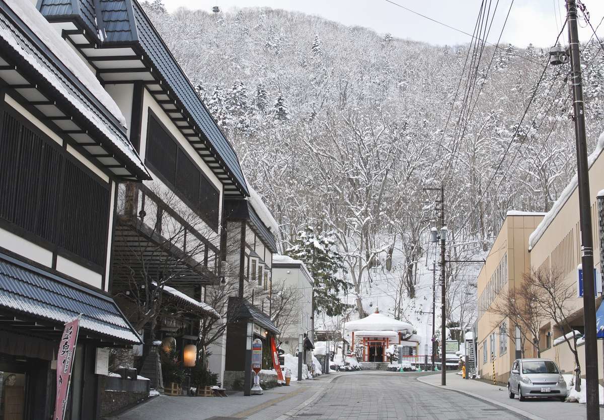 冬天白天的定山溪溫泉街:石板路左側是黑白相間的和風建築與燈籠,路的盡頭是一座屋頂積雪的紅色小堂,背後是整片雪林山坡