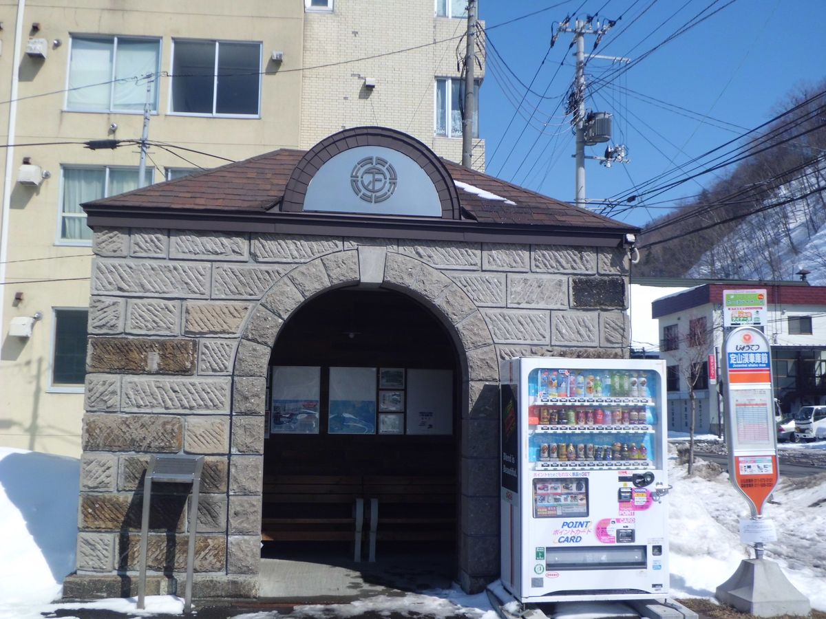 じょうてつ巴士「定山渓車庫前」站牌與石造拱門的候車亭,旁邊一台自動販賣機,地上有殘雪