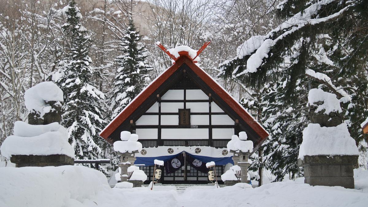 大雪中的定山溪神社:紅色屋頂、白牆黑框的社殿前掛著藍白布幕,兩側的石燈籠和狛犬都被雪埋住