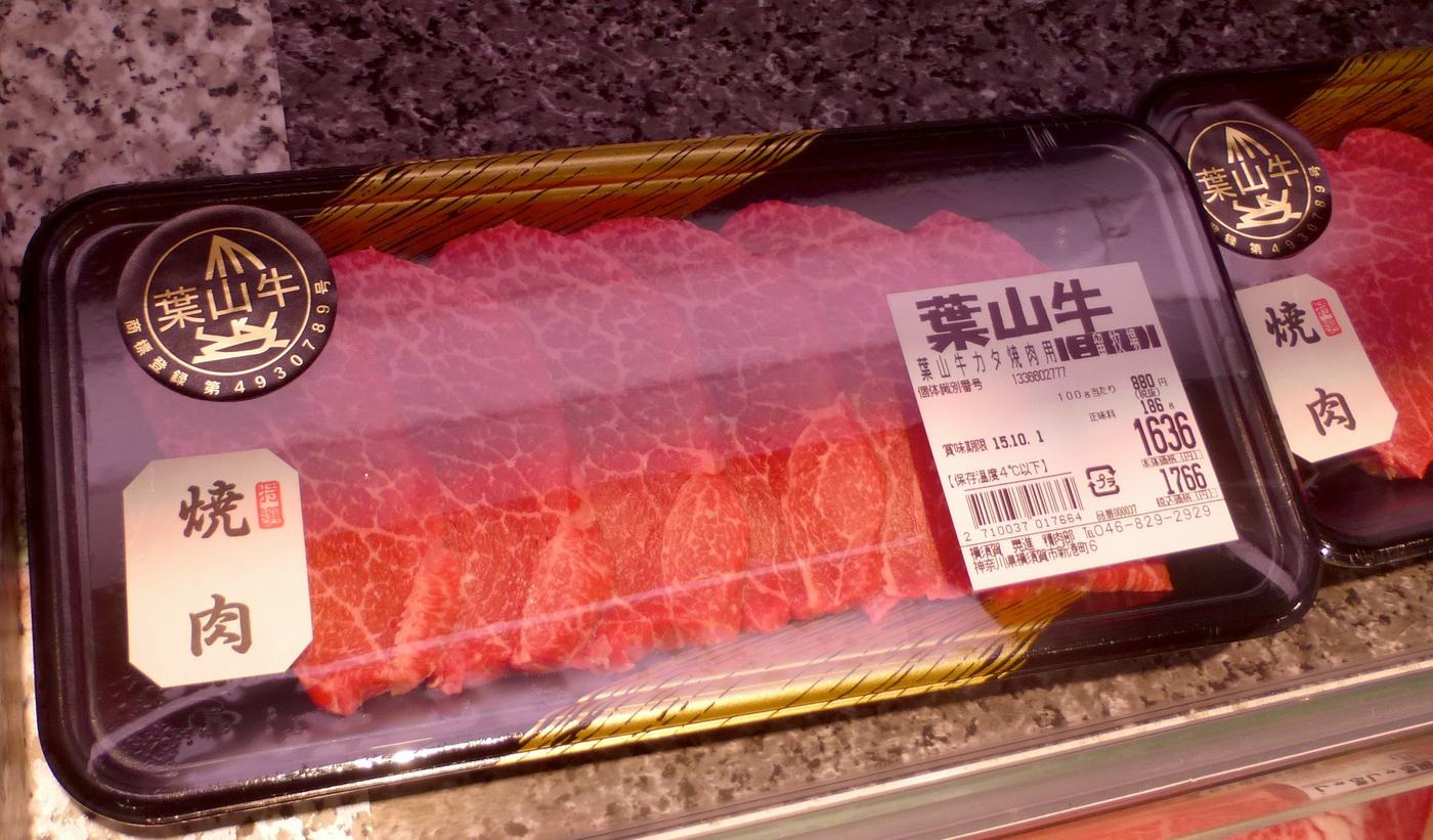 日本超市冷藏櫃裡的兩盤保鮮膜包裝生牛肉,黑色塑膠盤上貼著金色圓形「葉山牛」認證標與白底毛筆字的「焼肉」標籤,右側價格貼紙印著葉山牛カタ焼肉用、個体識別番号、100g 當 880 円、正味量 186g、税別 1636 円、税込 1766 円與賞味期限