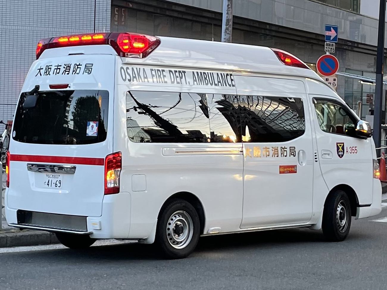 白色日本救護車停在市區道路上,車頂紅色警示燈亮著,車身上方以黑字寫著 OSAKA FIRE DEPT. AMBULANCE,側門與車尾上方另有「大阪市消防局」字樣,車側有紅色橫條與編號 A355