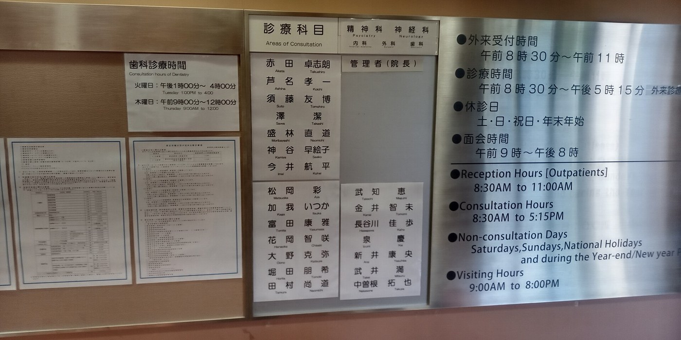 日本醫院牆上的揭示板:中央玻璃框內是「診療科目 Areas of Consultation」與一整排醫師姓名,右側不鏽鋼板上分中英文列出外來受付時間 8:30–11:00、診療時間 8:30–17:15、休診日與面會時間