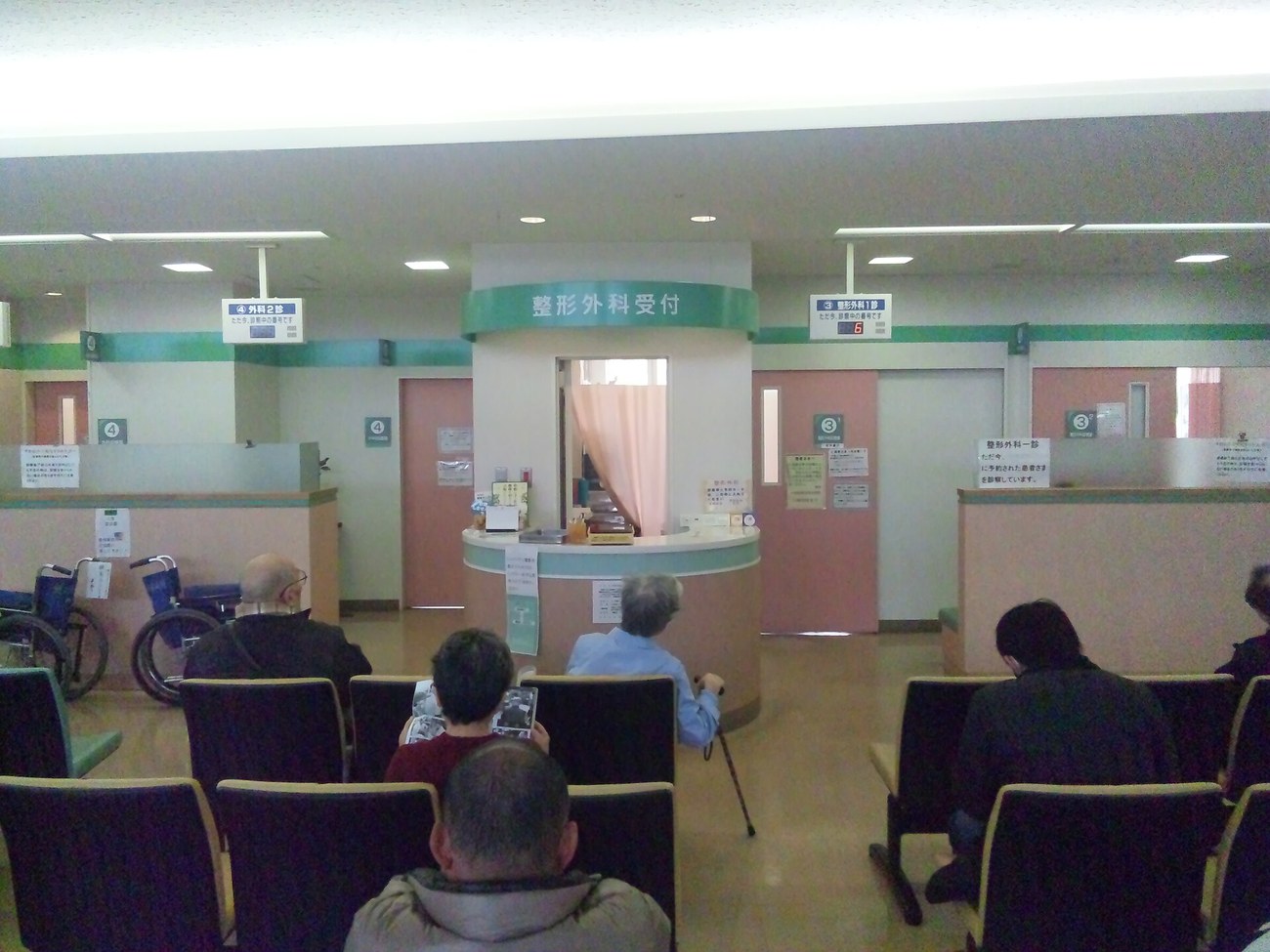 日本醫院候診區,中央是綠色圓弧櫃檯、上方掛著「整形外科受付」,左右兩側藍色吊牌分別寫著「④外科2診」與「③整形外科1診」並附紅色數字顯示器,前方成排深色座椅坐著候診的人,左邊停著兩台輪椅