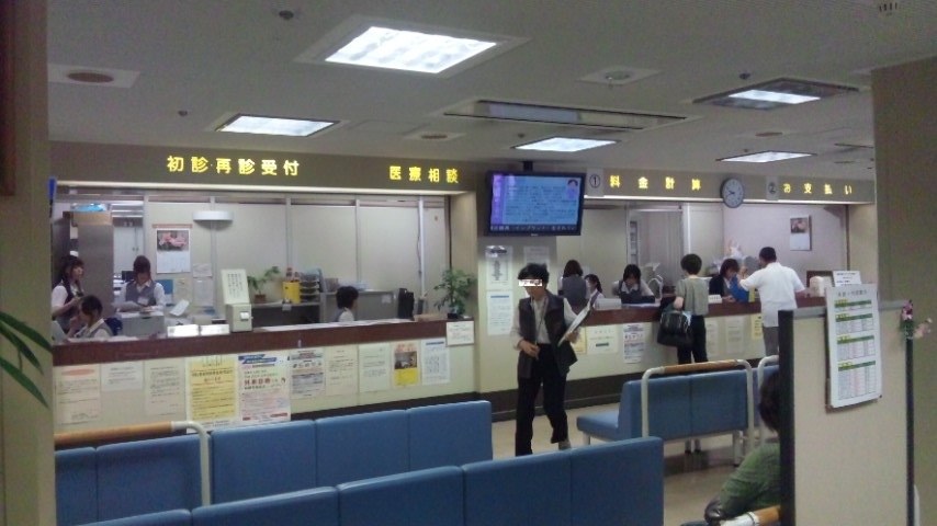 日本醫院的受付與會計區,深色橫樑上以黃字依序寫著「初診・再診受付」「医療相談」「料金計算」「お支払い」,中間掛著叫號螢幕與圓形時鐘,前方是成排藍色候診椅