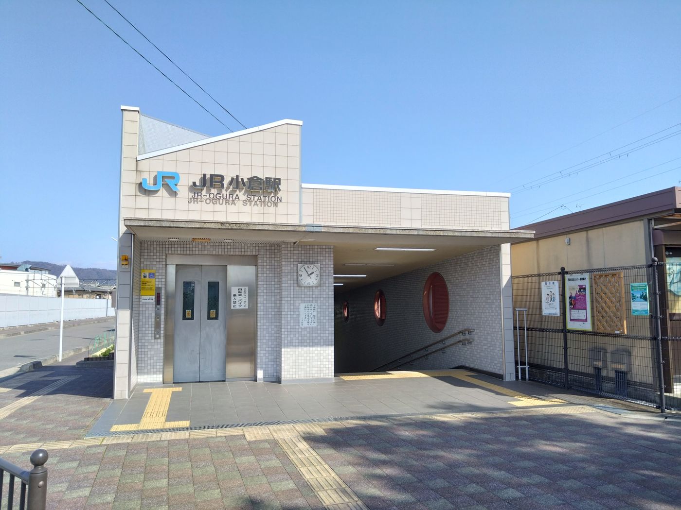 晴朗藍天下的小型車站南口:米色磁磚立面上是藍色 JR 標誌與黑字站名「JR 小倉駅」,底下一行英文「JR-OGURA STATION」;正面有一座銀色電梯門與一個圓形時鐘,右側是通往地下道的下行樓梯口,牆面嵌著三個大小不一的紅色圓形裝飾;站前是灰色磚鋪面與黃色導盲磚,畫面右邊是黑色柵欄與貼著海報的佈告欄,遠方可見低矮民宅與淺色山稜,站前無人