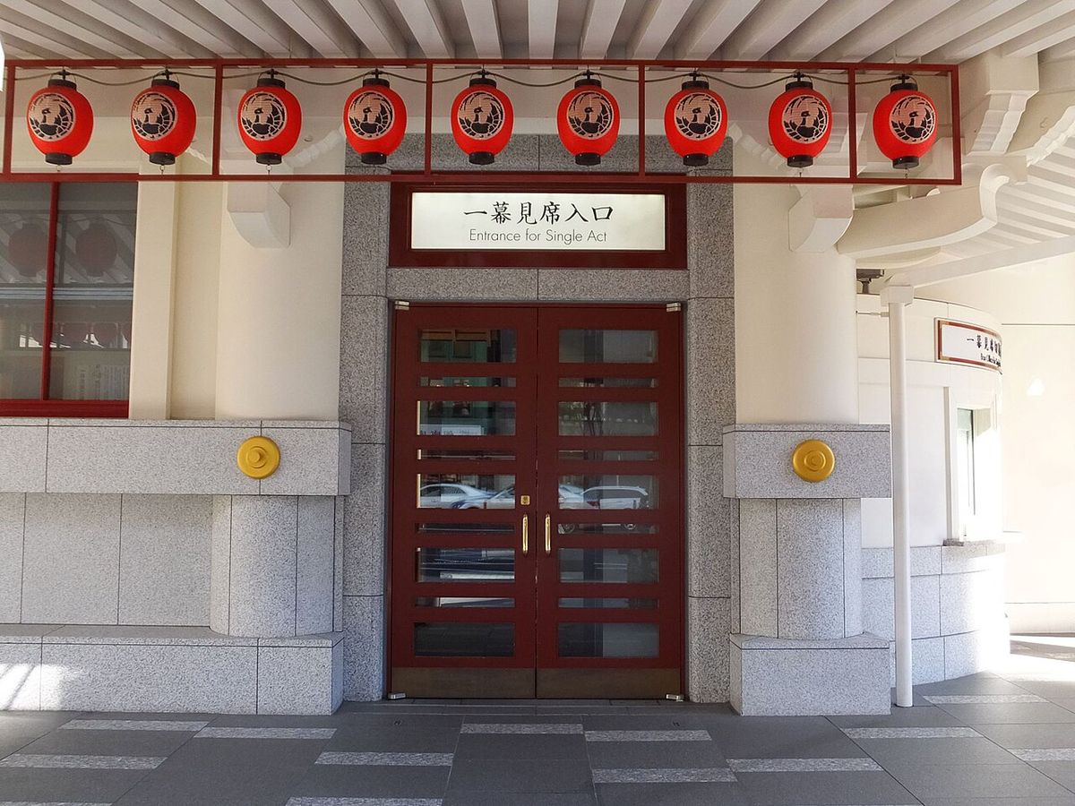 歌舞伎座一幕見席專用入口:暗紅色雙開玻璃門上方掛著白底黑字的「一幕見席入口 Entrance for Single Act」標示牌,再上方是一整排印有鳳凰丸座紋的紅色提燈,右側可見弧形白牆的售票亭一角