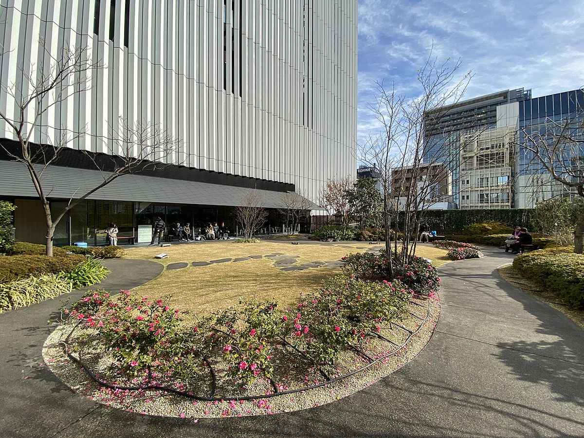 歌舞伎座五樓屋上庭園冬景:中央是枯黃的草坪與踏石步道,前景弧形花圃開著粉紅色山茶花,落葉期的樹枝光禿,左側玻璃帷幕店面前有人坐在長凳上,正上方是歌舞伎座塔的垂直白色格柵外牆