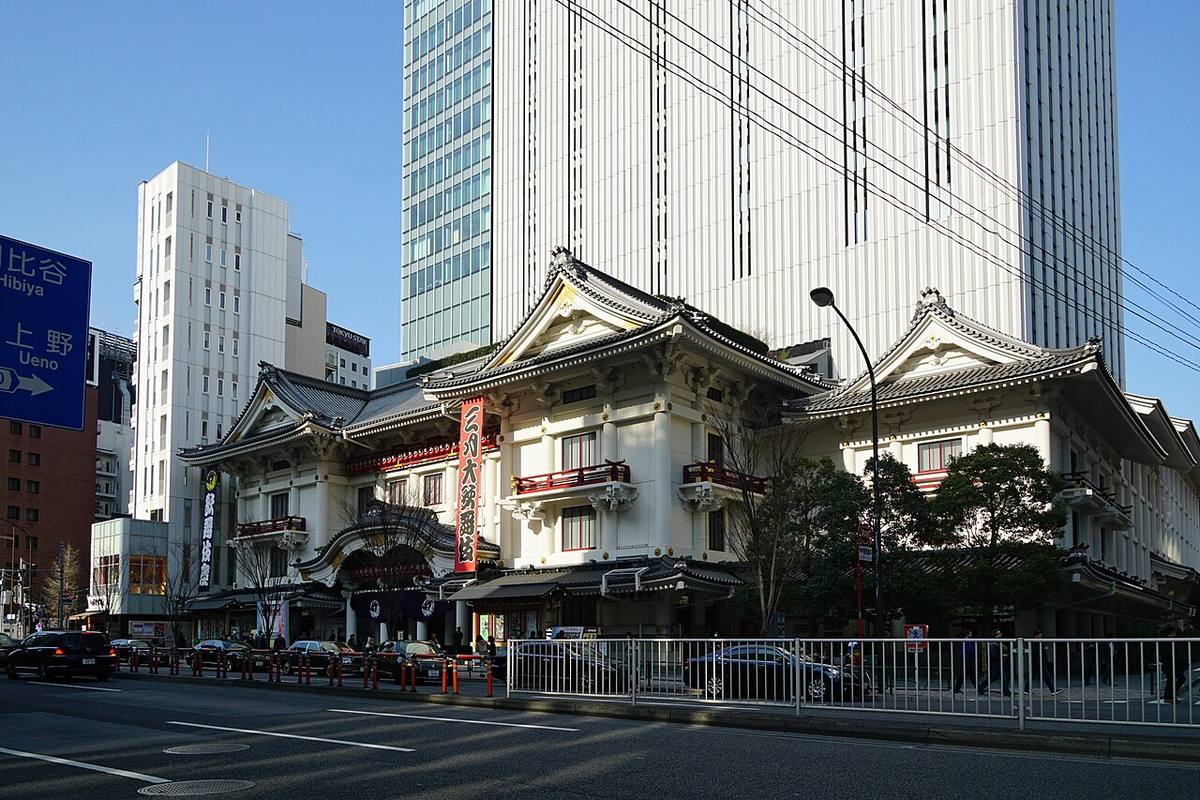 從晴海通り對街斜看歌舞伎座:三座白色破風屋頂橫向並排,中央掛著紅色直式公演看板,左端有深色直式布幔,背後是灰色的歌舞伎座塔,路口立著往日比谷、上野方向的藍色道路指示牌