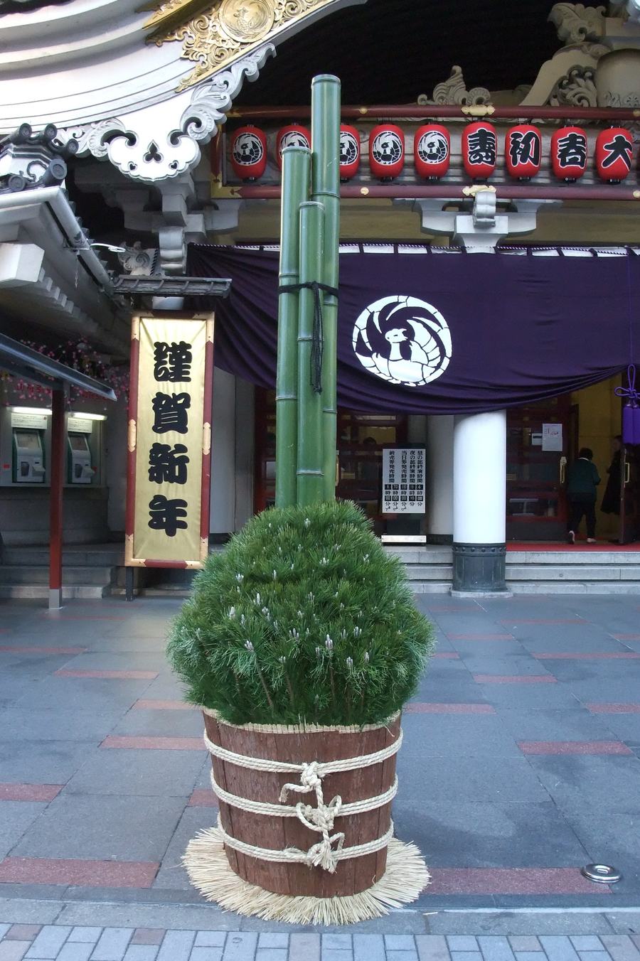 歌舞伎座正面的大型門松,三根斜切青竹立於松枝與草繩綁縛的木桶中,後方掛著謹賀新年立牌與紅色提燈