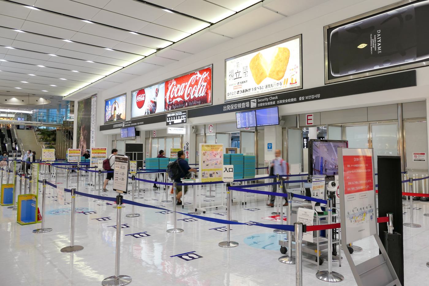 鹿兒島機場出發保安檢查場,天花板下方的黑底看板寫著「出発保安検査場 Security Check」並附簡體中文、繁體中文與韓文,右側標著 B、C、E 通道編號,地面以藍色伸縮帶圍出排隊動線,檢查台旁堆著成落的青綠色托盤