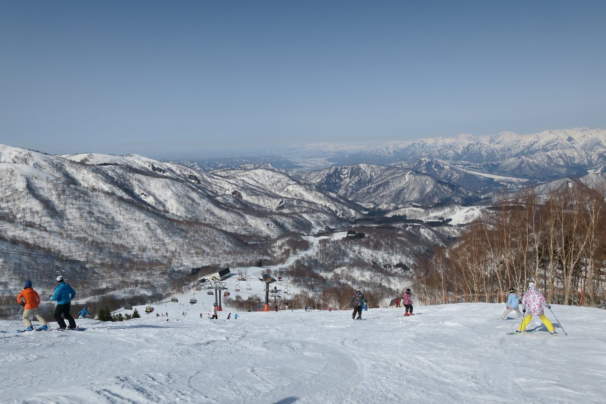 かぐら滑雪場上方的雪道：幾名滑雪者背對鏡頭往下滑，下方山谷有吊椅纜車和山屋，遠方是一層層覆雪的山脈，更遠處是平原