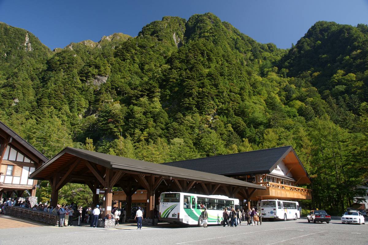 上高地巴士總站木造月台前停靠的接駁巴士與排隊旅客，背後是陡峭山壁