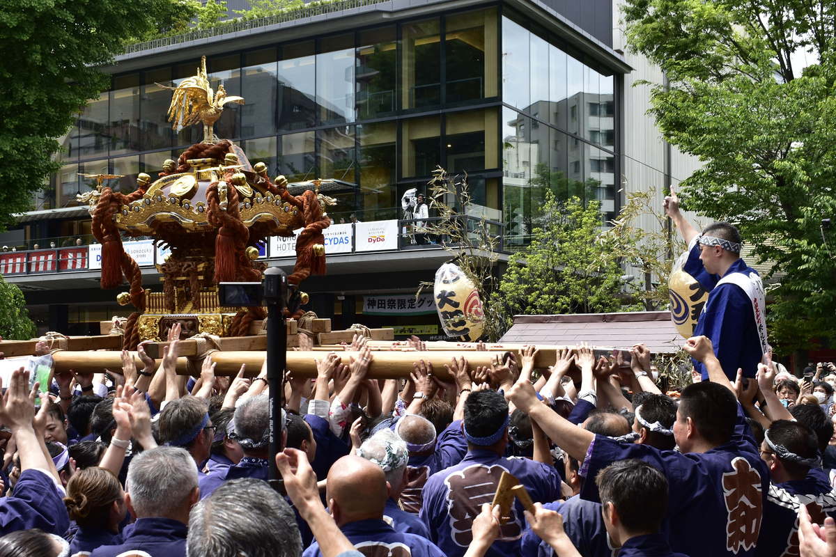 神田祭神輿被高高抬起：金色神輿頂著金鳳凰、周身纏著粗麻繩，橫木上密密麻麻全是舉起的手，抬轎者身穿藍染半纏，右側一名男子站在高處舉手指揮，背景是神田明神文化交流館 EDOCCO 的玻璃帷幕與「神田祭限定グッズ」布條